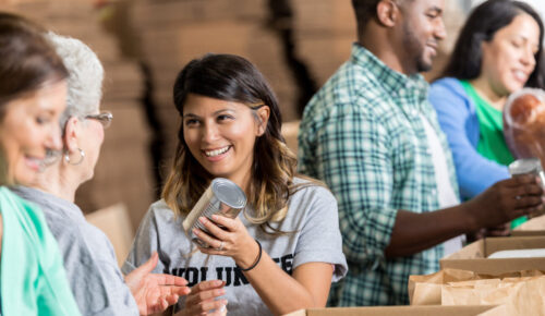 Beautiful mid adult Hispanic woman accepts a donation during a community food drive. She and her friends are packing cardboard boxes. Diverse people are also volunteering in the background.